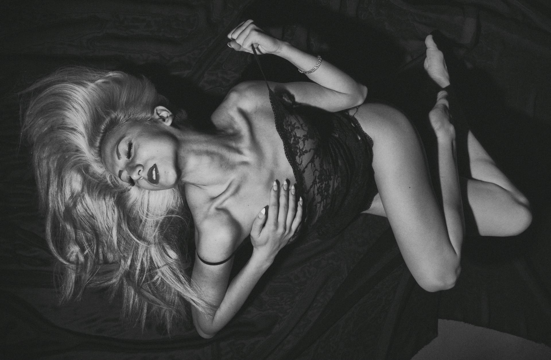 Black-and-white lifestyle photo of woman in lace bodysuit reclining on dark fabric, NipLift